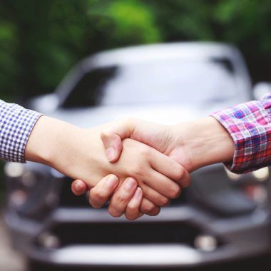 A man shakes hands with another man. A car is in the background