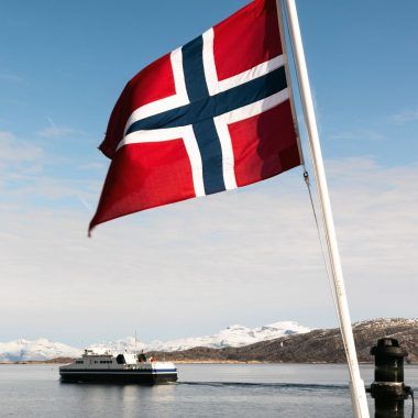 Norwegian flag flying from a boat. Another boat and fjords in the background.
